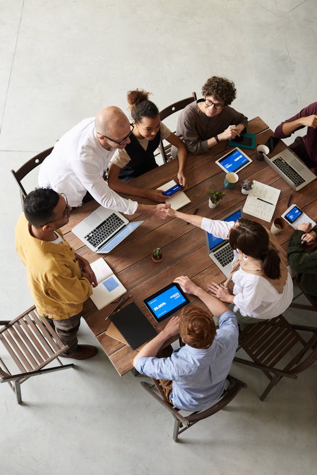 A diverse group of eight colleagues sit around a wooden table with laptops and tablets, engaged in discussion.